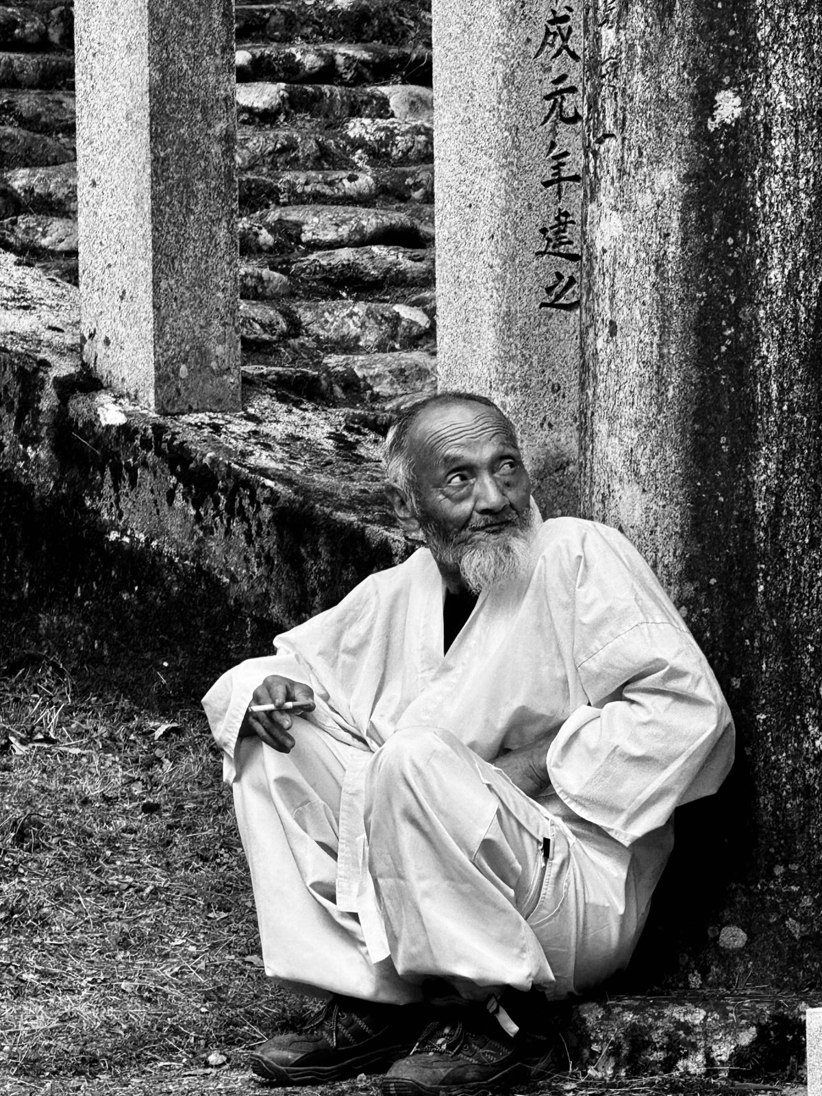 Old man in white sitting against stone pillars, black and white